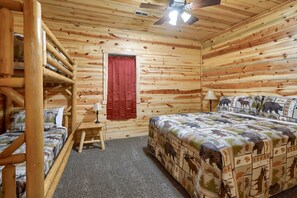 A log cabin room with a bed, a small table, a lamp, a ceiling fan, and a red curtain. - Waccabuck | 17.5 Private Acres, Pool, Hot Tub (Logan)