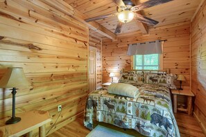 A wooden cabin room with a bed, two lamps, a ceiling fan, and a window with curtains.