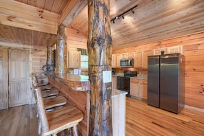 A rustic cabin interior with a wooden bar, seating, a large central tree trunk, and a stainless steel refrigerator.