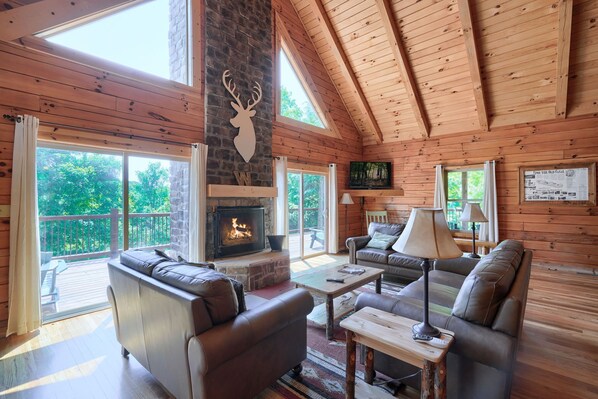 A cozy cabin interior with a fireplace, leather sofas, a television, and a large window overlooking greenery. - Waccabuck | 17.5 Private Acres, Pool, Hot Tub (Logan)