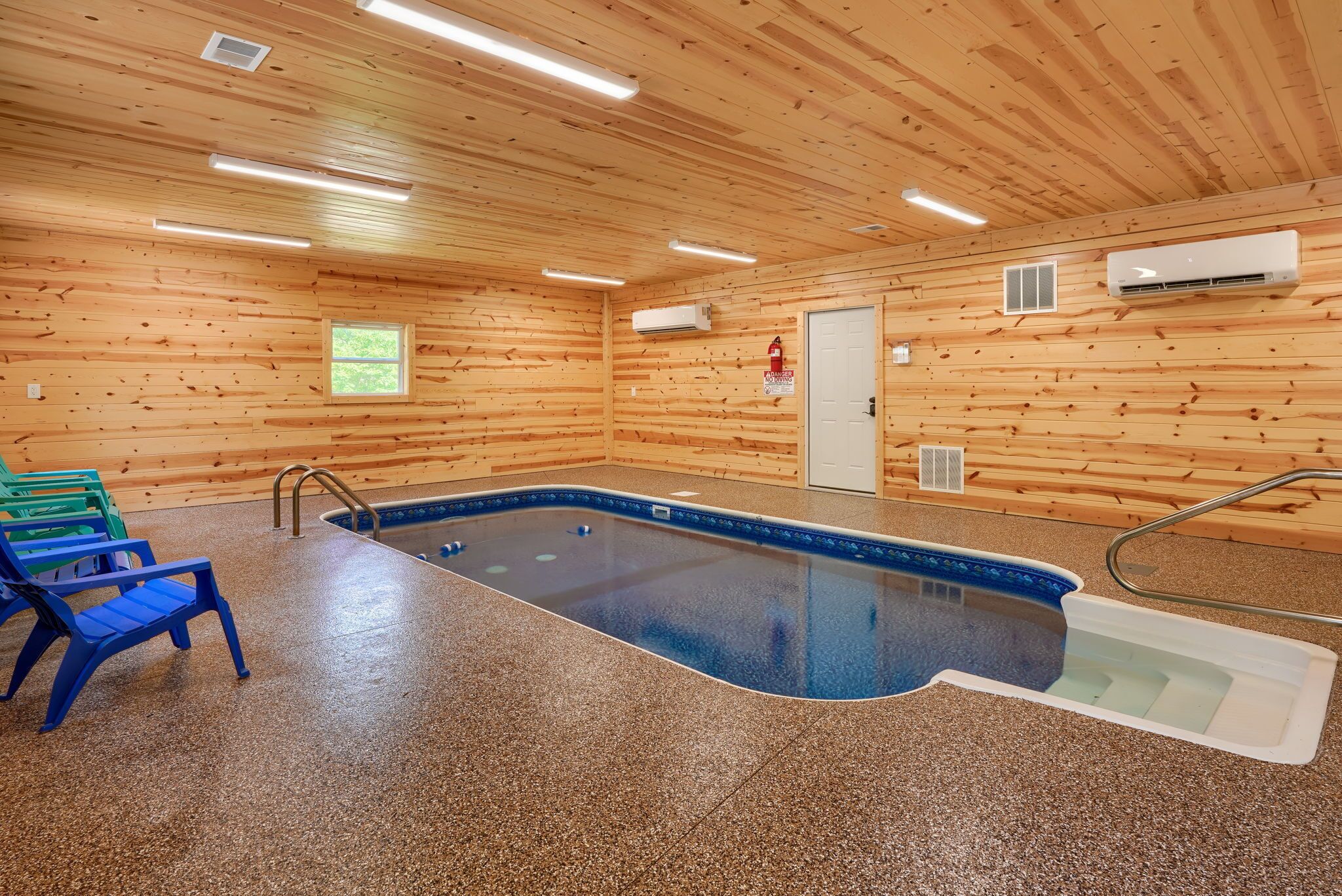 An indoor swimming pool with a blue border, surrounded by a pebble-textured floor, wooden walls, and a door with a fire extinguisher.