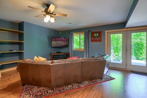 A living room with a brown leather sofa, a ceiling fan, a television, and a large window with a view of greenery. - Sandstone Valley Estate | Hocking, Indoor Pool (Logan)