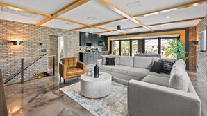 A modern living room with a large grey sofa, a round coffee table, and a leather armchair. A brick wall and wooden beams are visible in the background.