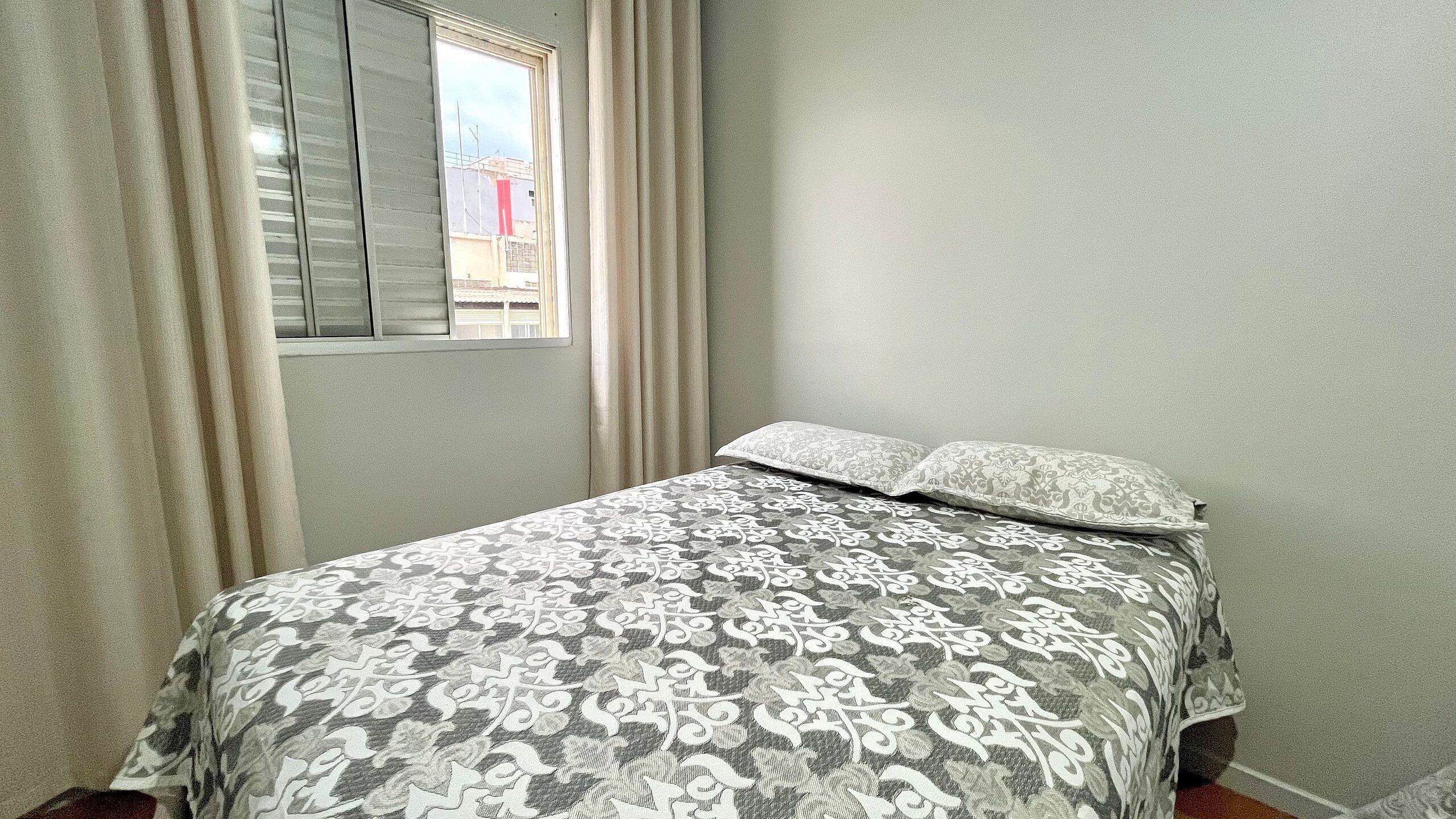 Un dormitori amb un llit, cortines, una finestra i una paret sense decorar.
