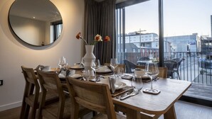 Un comedor con una mesa redonda, sillas, un florero con flores y un ventanal con vistas a edificios.