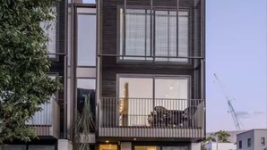 Un edificio residencial moderno de dos pisos con balcón, ventanas grandes y entrada visible.