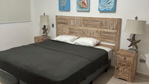 A bedroom with a wooden headboard, a bed with white pillows, and a dark bedspread. There are three wall-mounted paintings above the bed, and two nightstands with starfish lamps.
