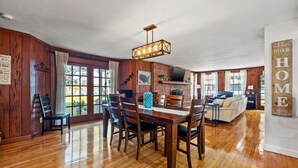 Un comedor amplio con una mesa y sillas de madera, una araña y vistas al exterior a través de grandes ventanales.