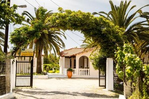 Une entrée fermée par un portail mène à un bâtiment doté d’un toit en tuiles, entouré d’une végétation luxuriante et de palmiers.