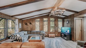 A cozy living room with a sectional sofa, leather ottomans, a television, and a large window offering a view of the outdoors. - Welcome to The Baker House Retreat - The Ranch House and The Bunk House (Maple Falls)