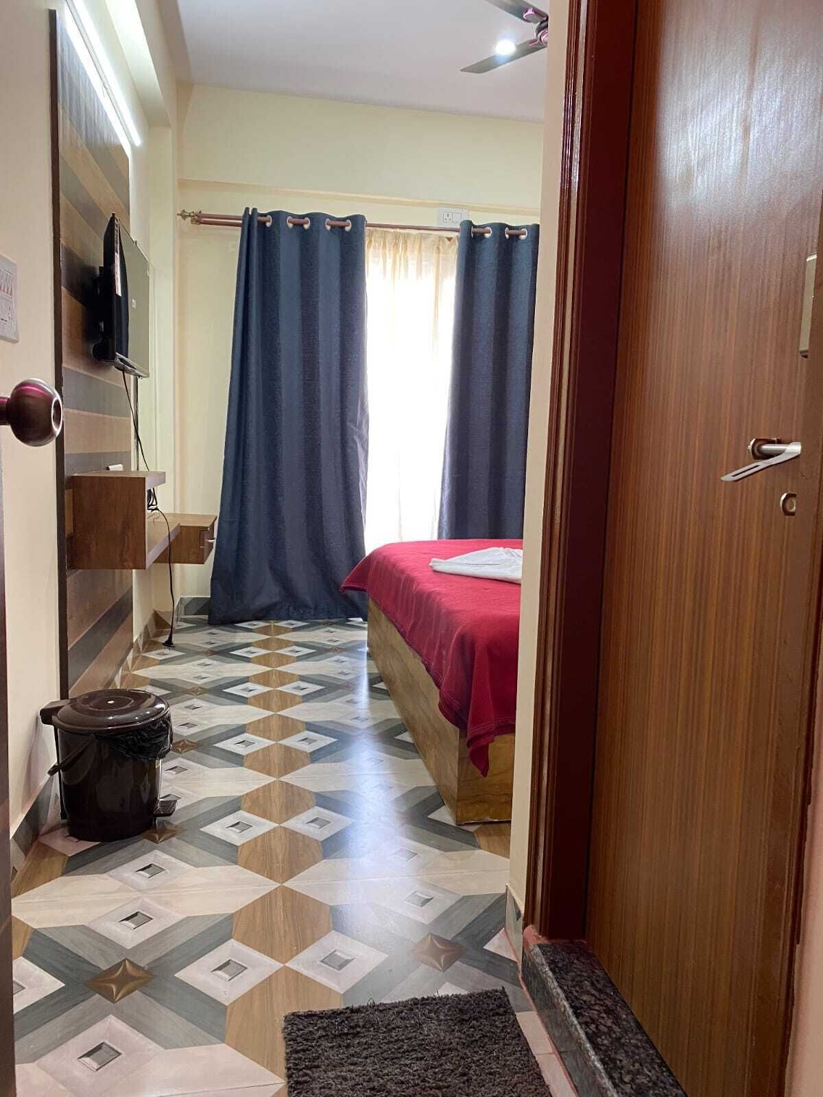 A hotel room with a bed, a TV mounted on the wall, a wooden wardrobe, a trash bin, and a patterned floor.