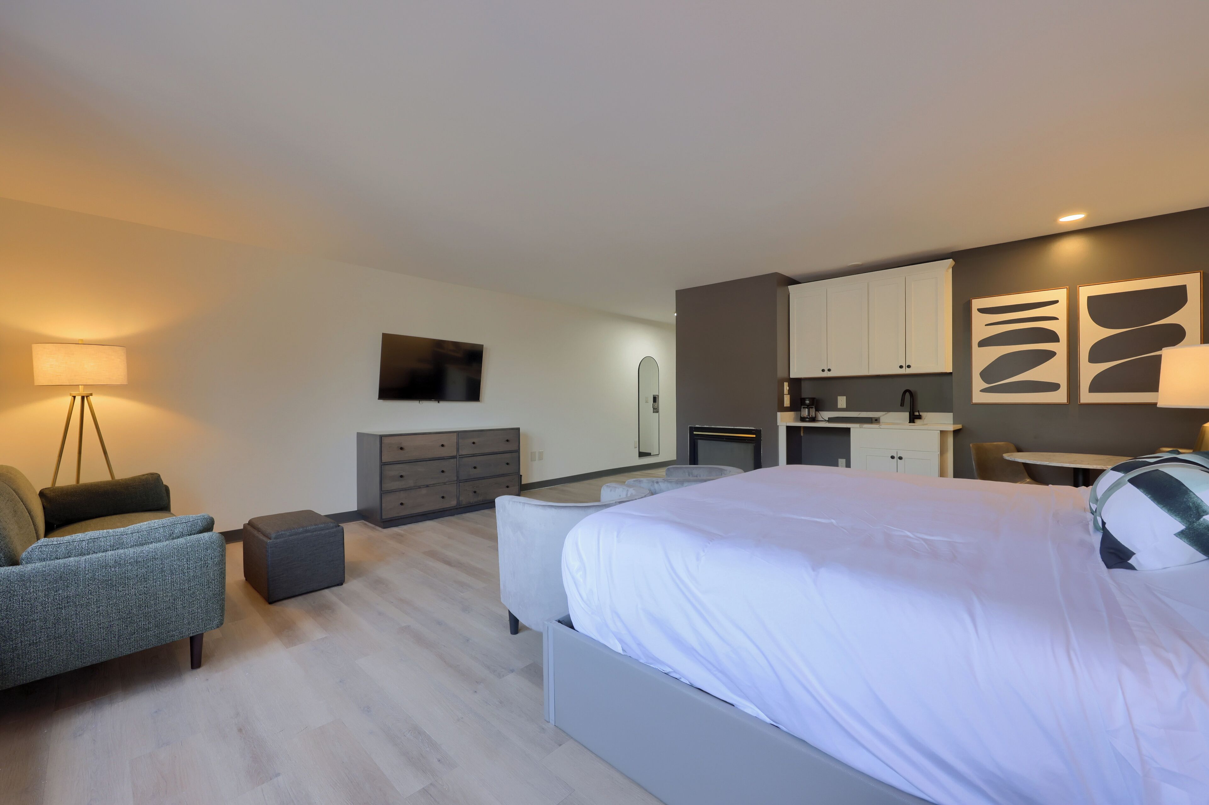 A modern hotel room with a large bed, a sofa, a desk, and a small kitchen area.