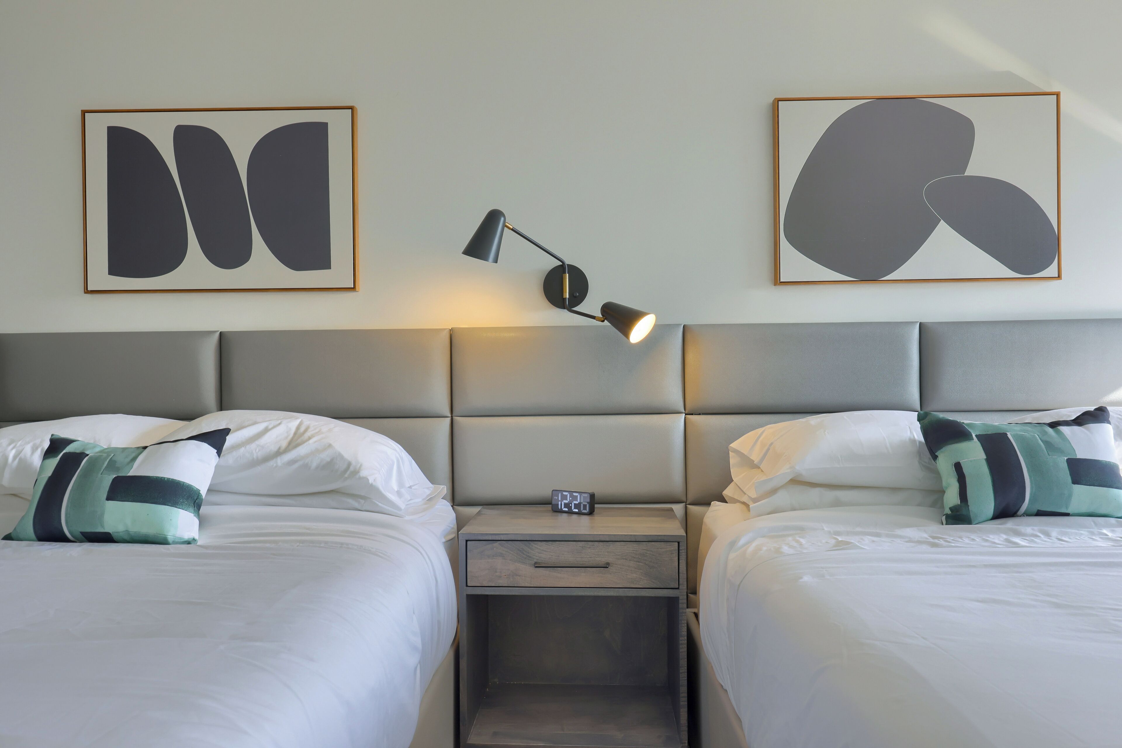 A hotel room with two beds, a nightstand with a clock, and abstract art on the wall.