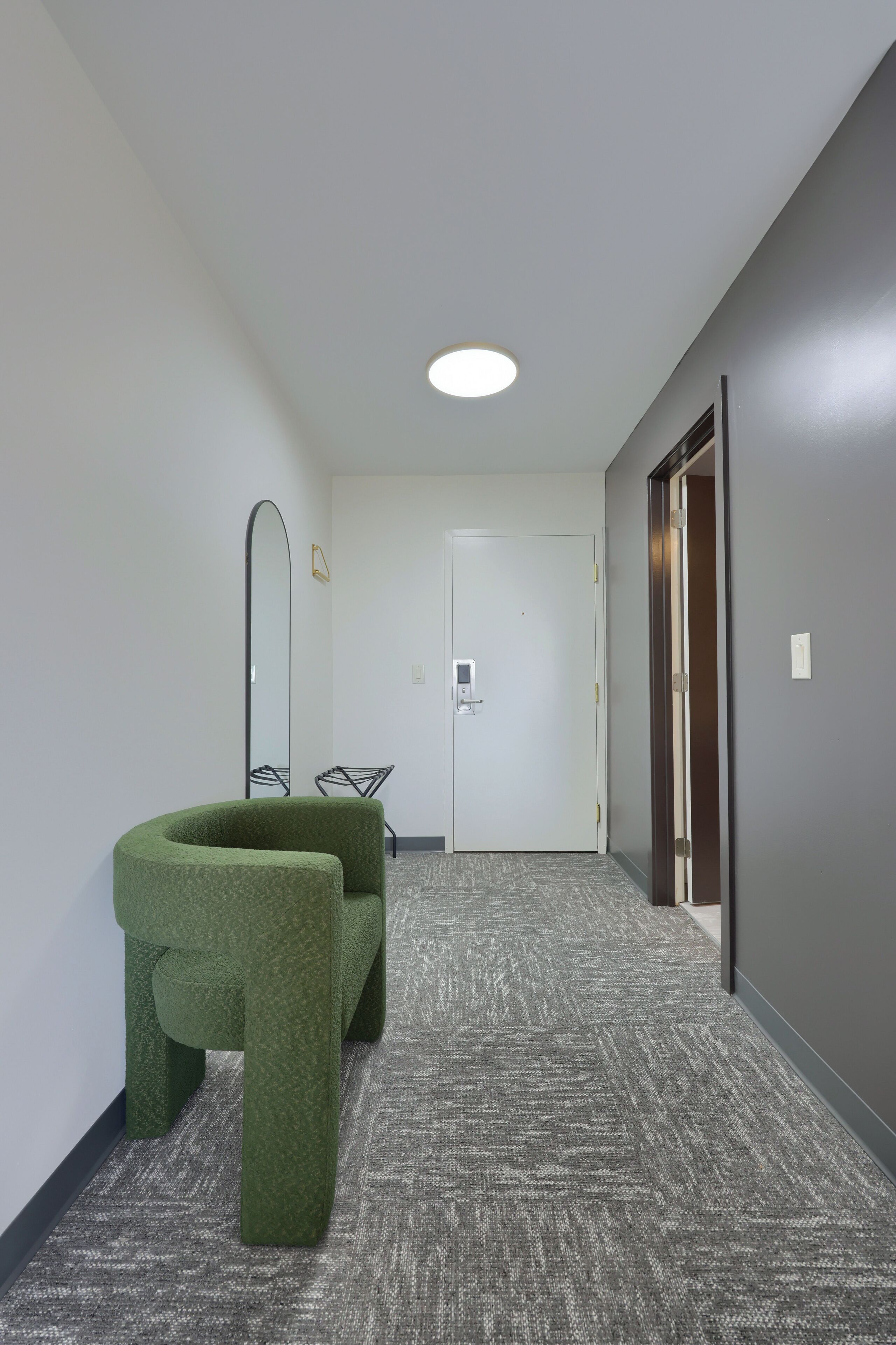 A hallway with a carpeted floor, a green upholstered chair, a mirror, and a door at the end.