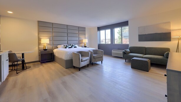 A modern hotel room with a large bed, two bedside tables with lamps, a grey sofa, a small ottoman, and a desk with a chair. - Lititz Pointe Inn and Suites (Lititz)
