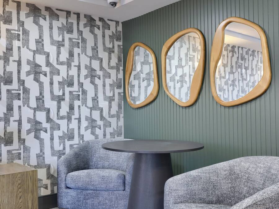 A room with a patterned wallpaper, two armchairs, a round table, and a mirror with a wooden frame.