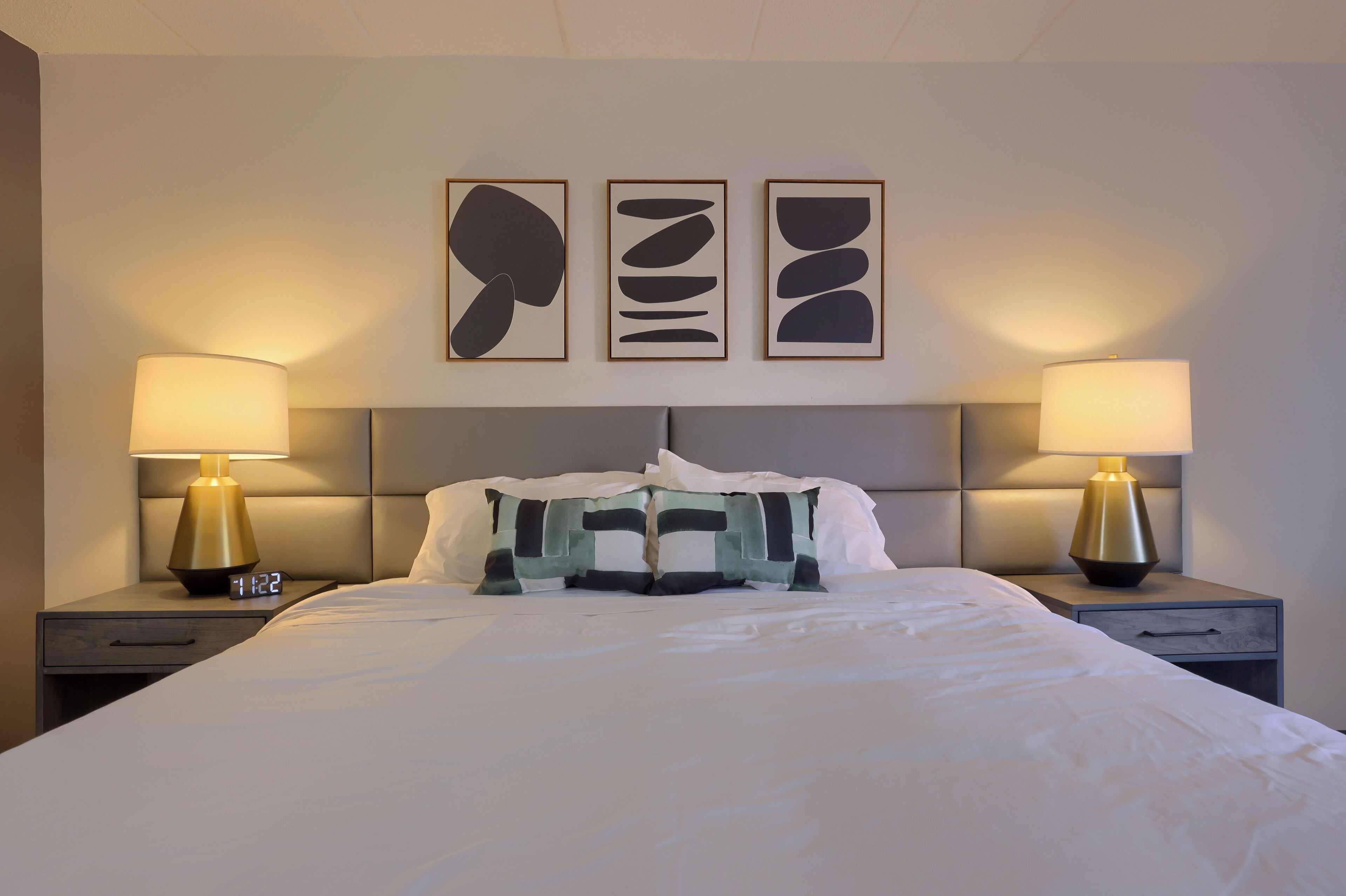 Una cama bien tendida con un cabecero, dos mesitas de noche con lámparas y tres obras de arte abstracto en la pared.