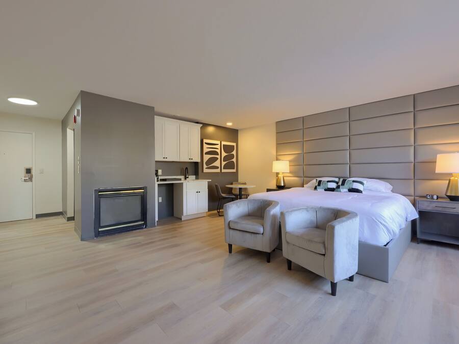 A modern hotel room with a bed, two armchairs, a small desk, and a built-in kitchen area.
