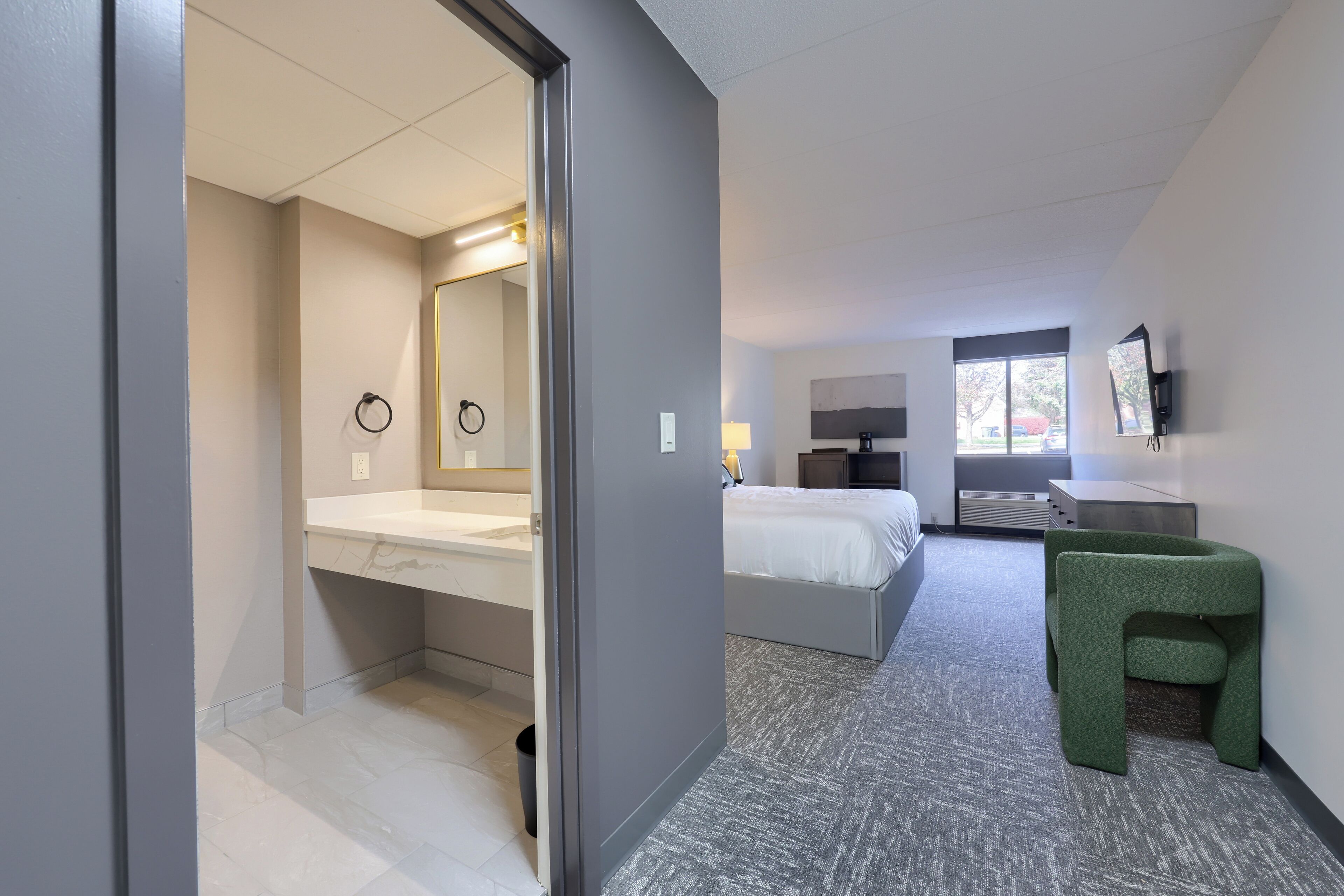 A modern hotel room with a large bed, a separate bathroom, a green upholstered chair, and a flat-screen TV mounted on the wall.
