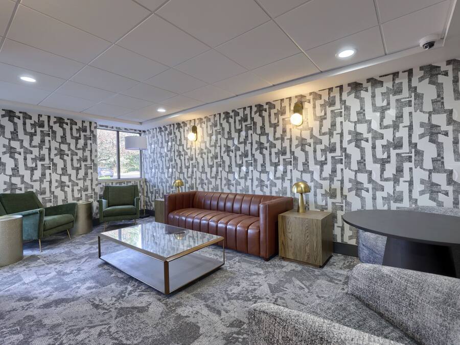 A modern lounge area with a brown leather sofa, green armchairs, a glass coffee table, and patterned wallpaper.
