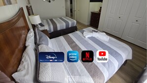 Habitació d'hotel amb llit, taula de nit, làmpada i paret amb miralls decoratius. El llençol té logotips de Disney+, Prime Video, Netflix i YouTube.