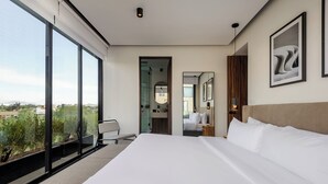 A modern hotel room with a large bed, a seating area with a bench, a view of the city through large windows, and a bathroom visible through an open door. - Zacatecas 4 by Wynwood House  (Mexico City)