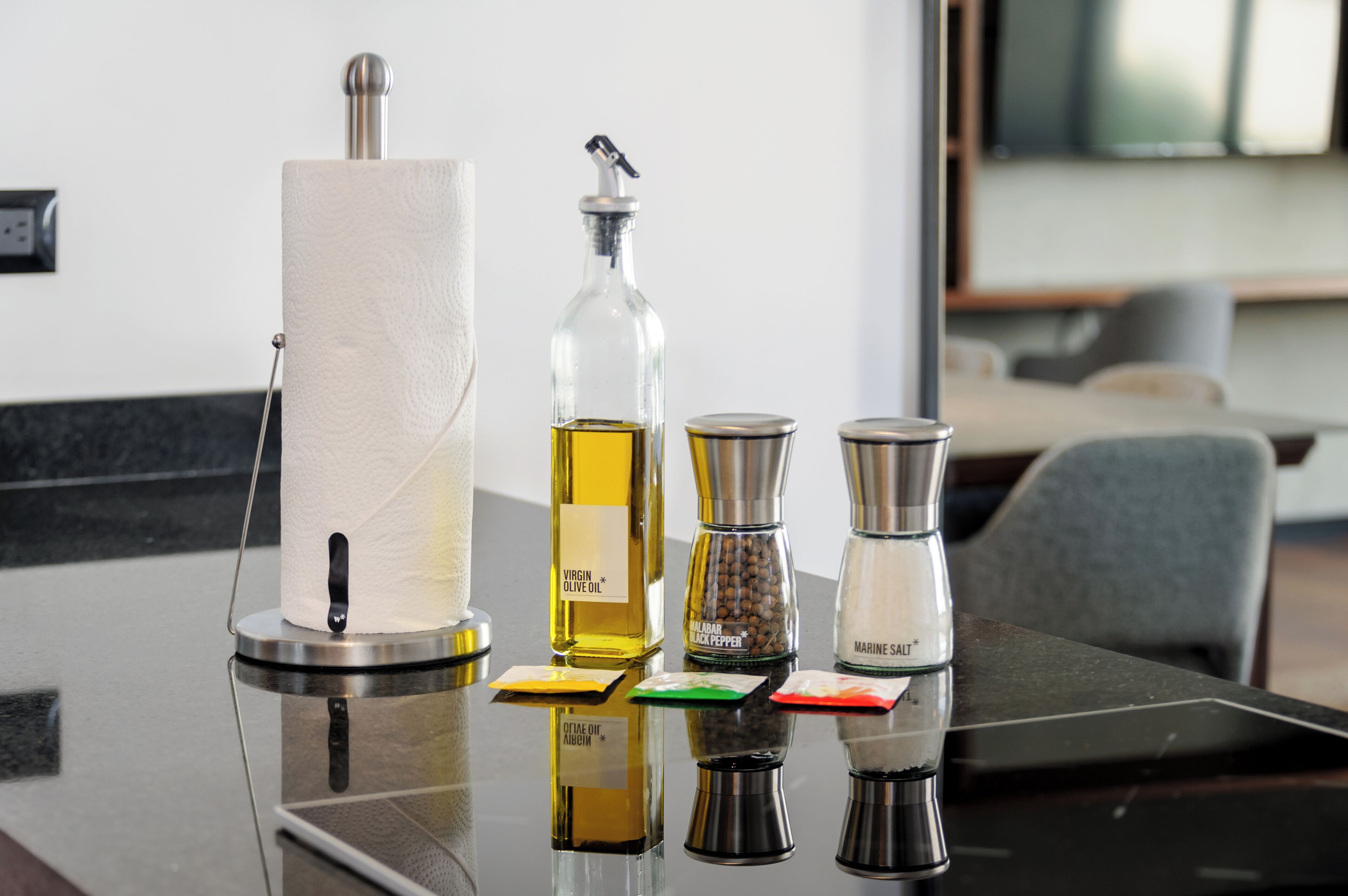 A table with a paper towel dispenser, a bottle of olive oil, a pepper grinder, and a salt shaker.