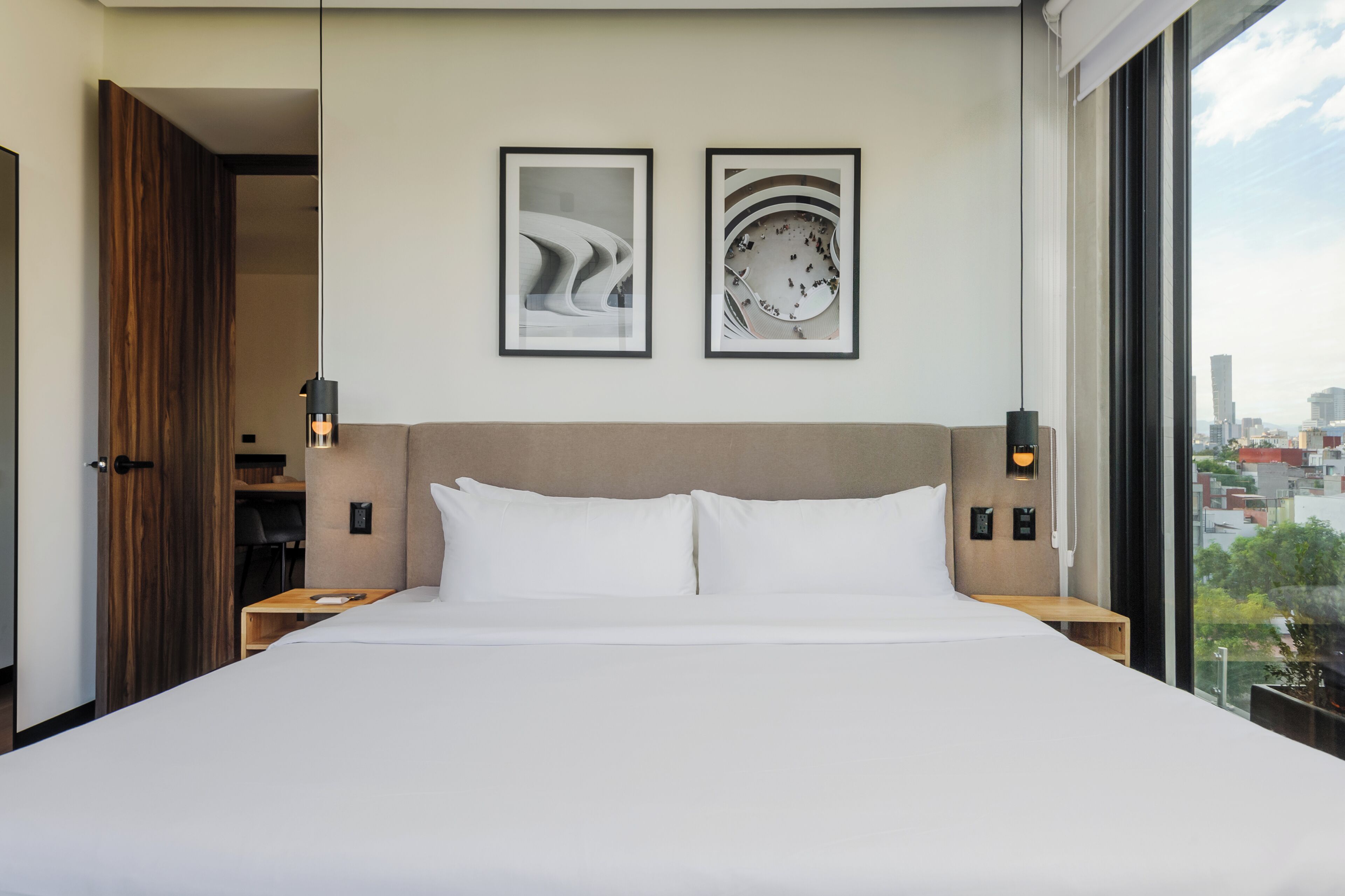 A modern hotel room with a large bed, two framed artworks on the wall, a city view through the window, and a wooden door.