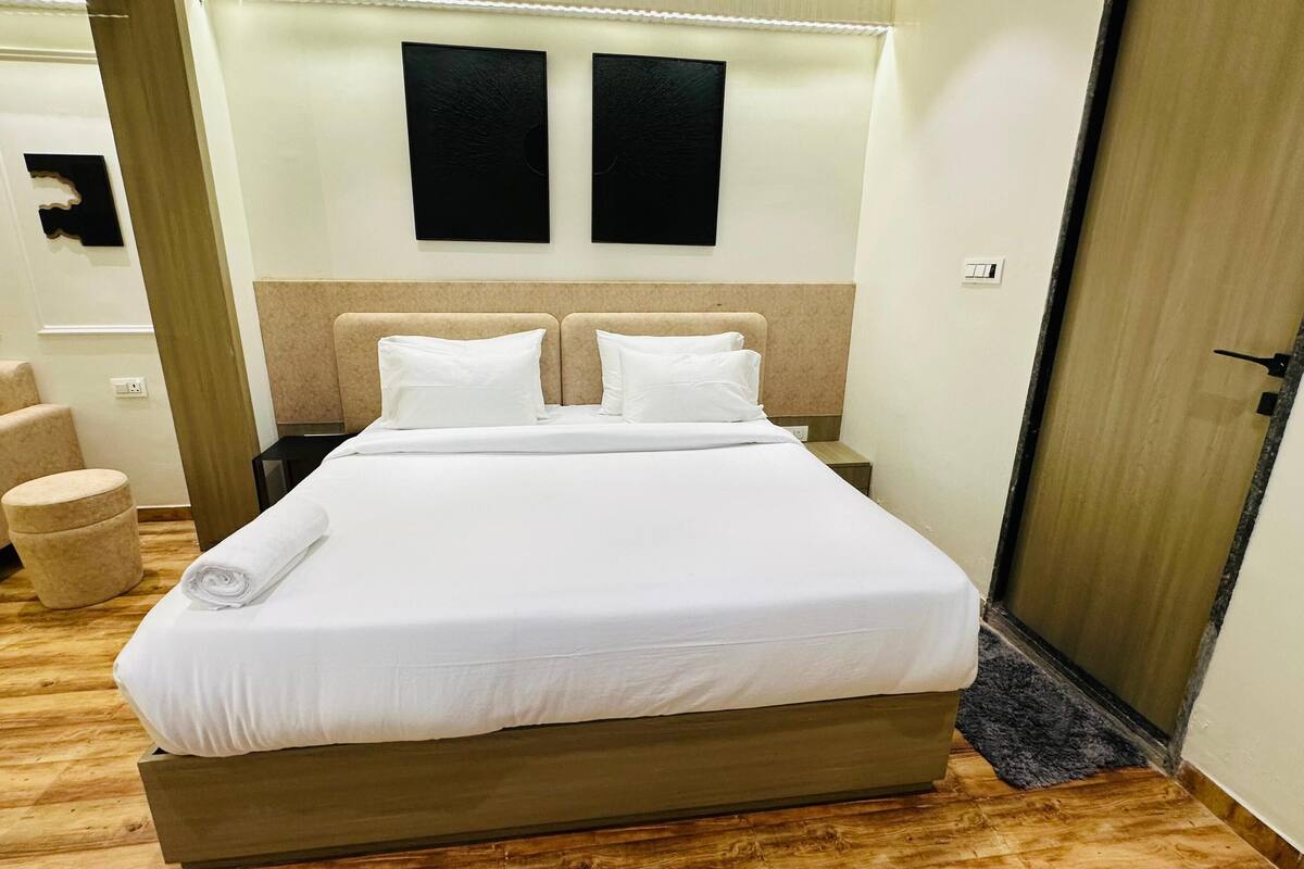 Habitación de hotel con una cama grande, cabecera, mesita de noche pequeña, una silla y un otoman redondo.