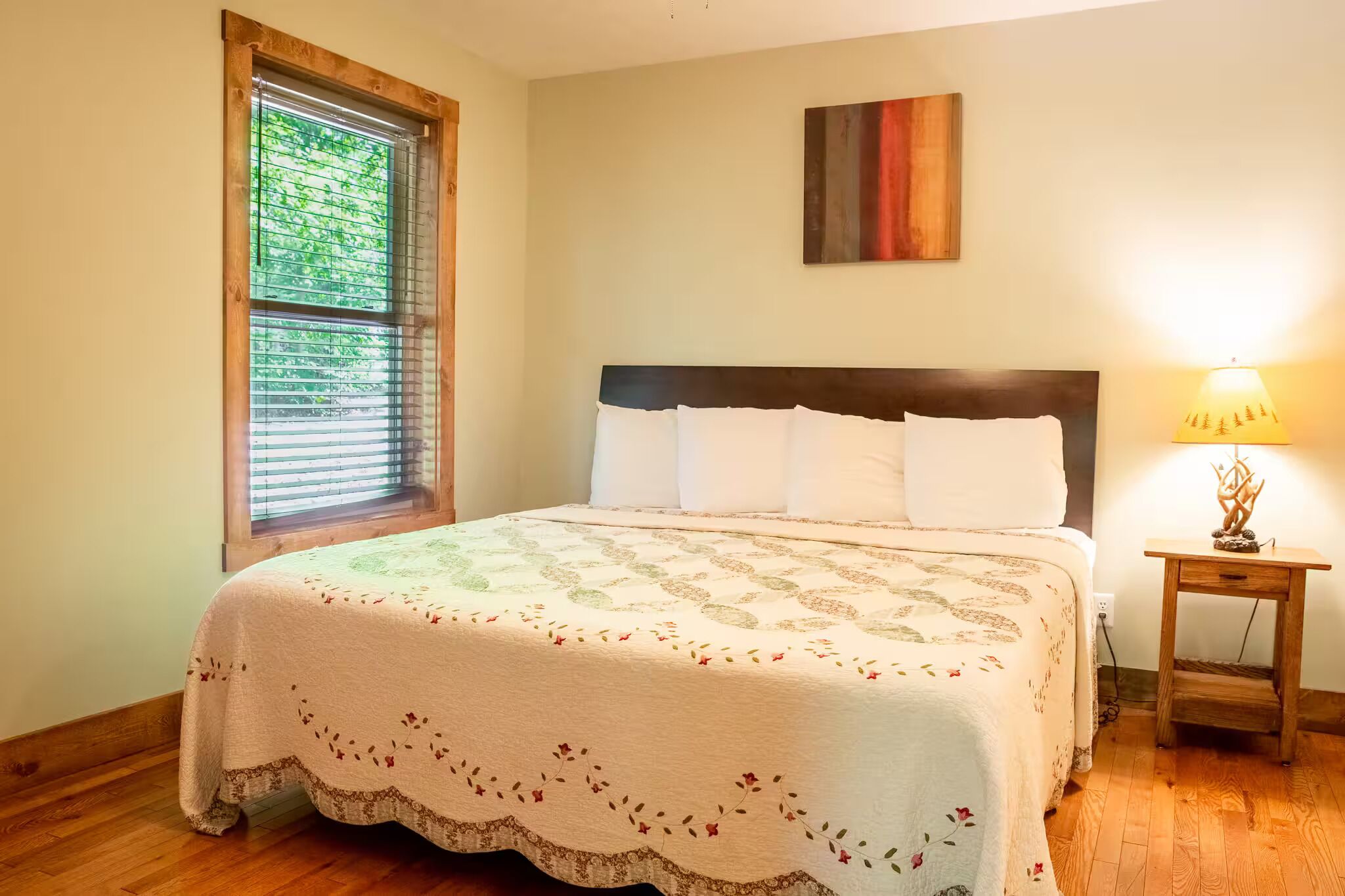 A bedroom with a bed, a window with blinds, a nightstand with a lamp, and a painting on the wall.