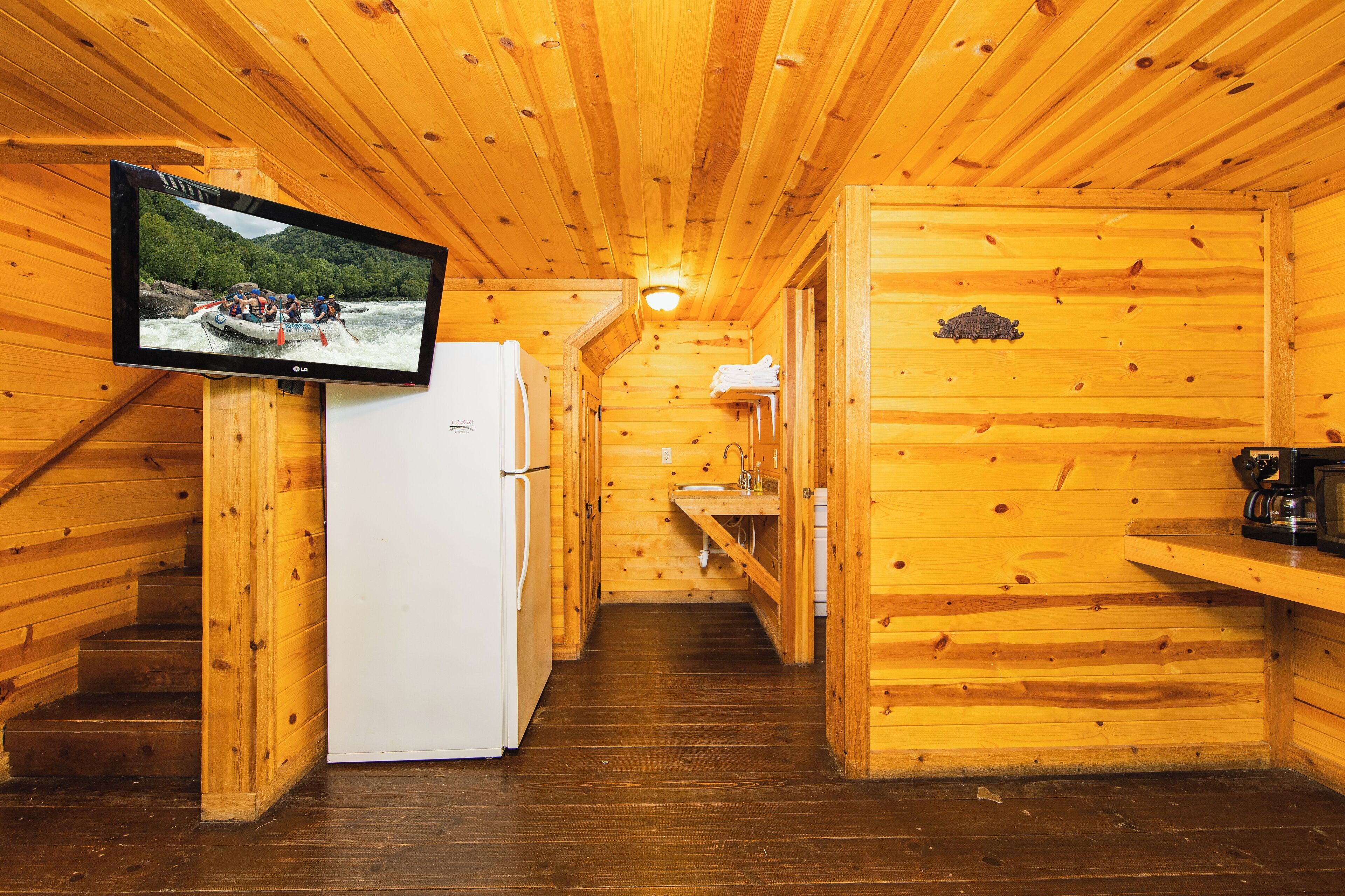 A wooden cabin interior with a kitchen area, a refrigerator, a television mounted on the wall, and a wooden staircase.