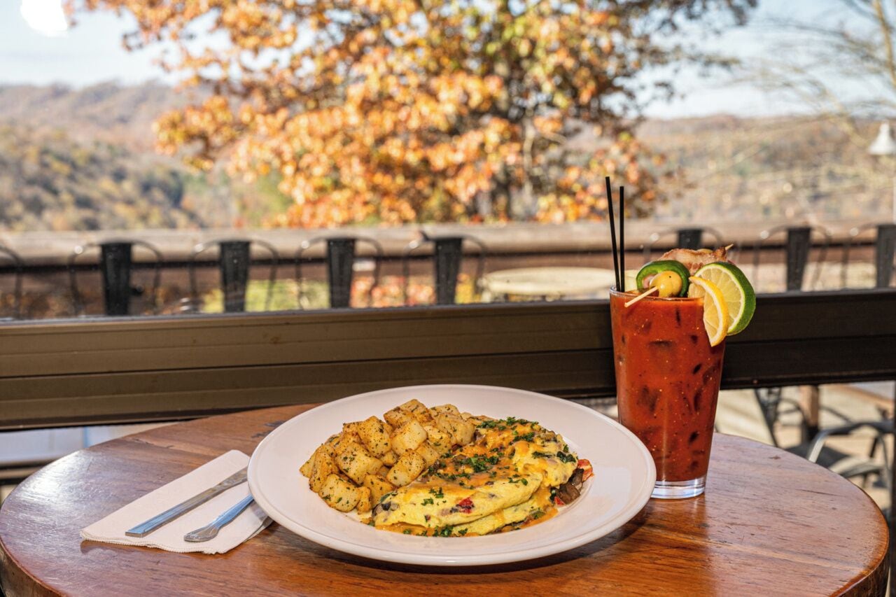 A meal with a plate of food and a drink on a table with a view of autumn trees.