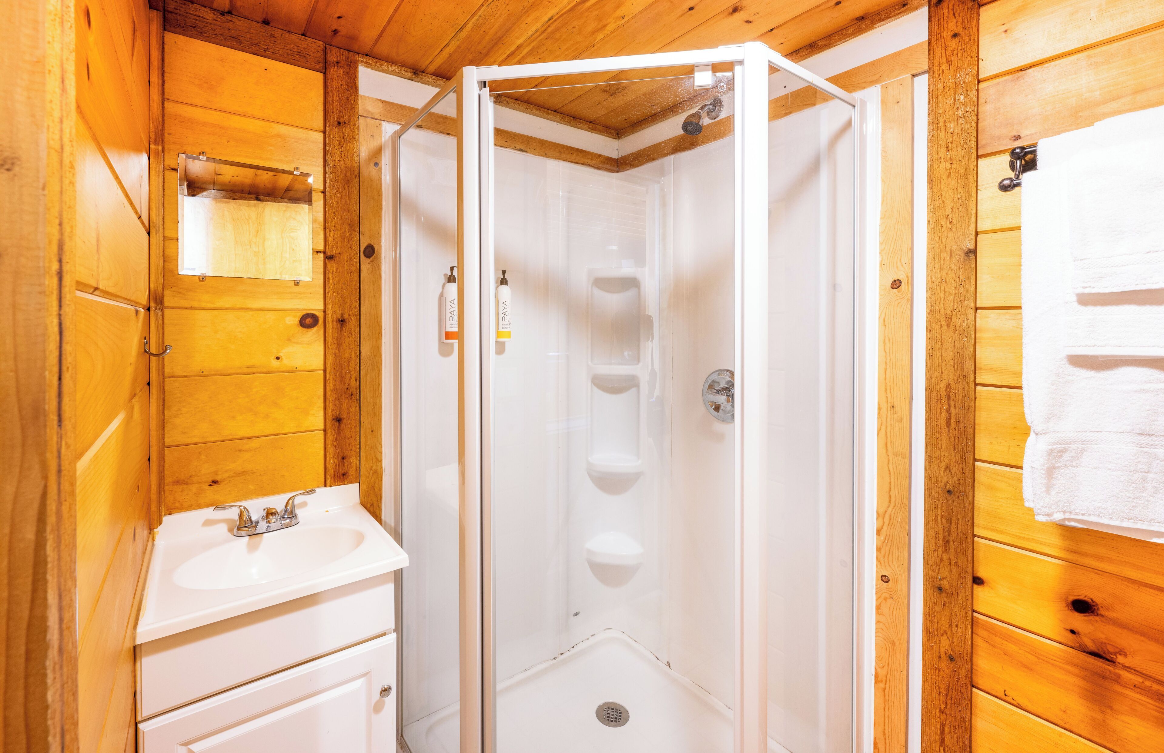 A wooden cabin bathroom with a white sink, a glass shower enclosure, and a towel hanging on the wall.