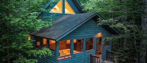 A cabin with a triangular roof, built into a forested area, featuring a deck and a sign that reads "Heaven Help Us."