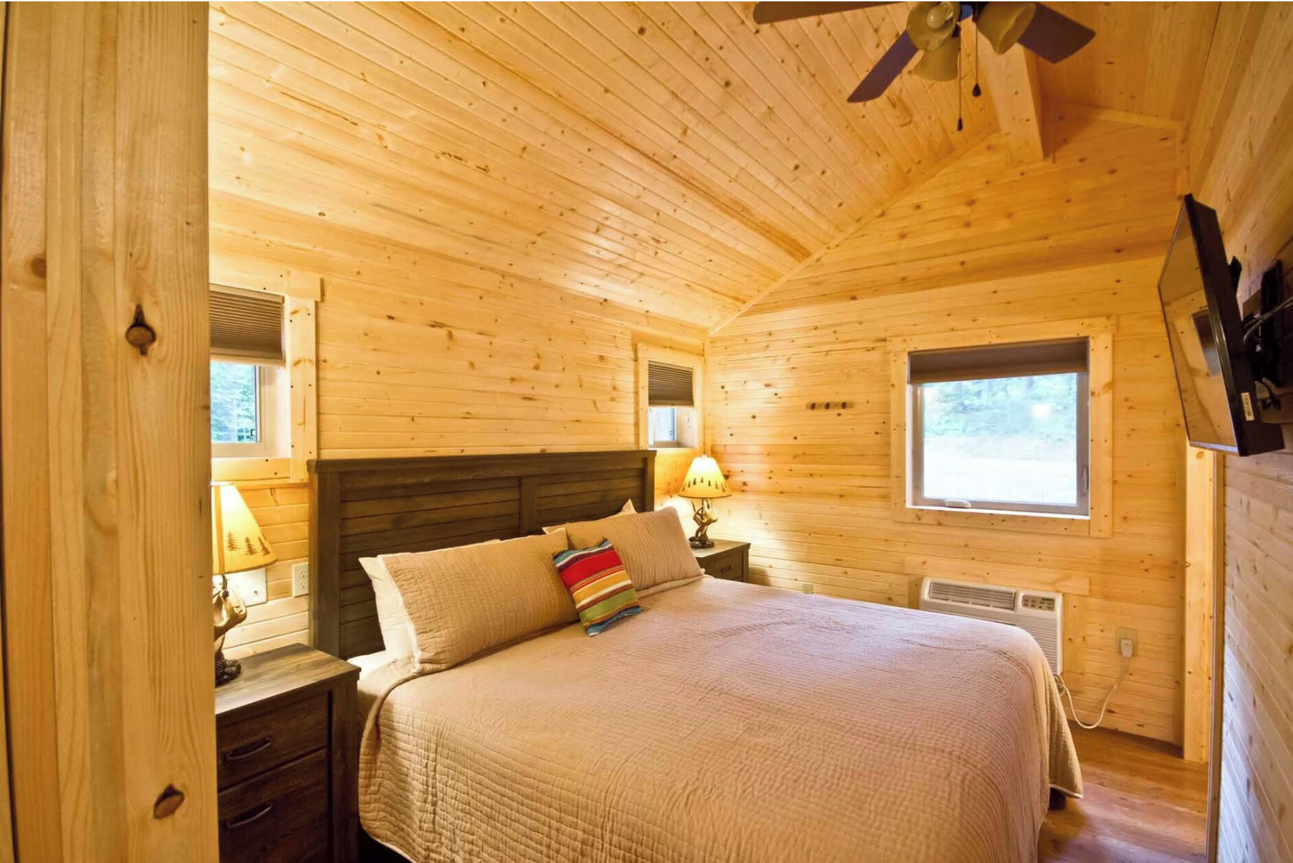 Un dormitorio acogedor de cabaña de madera con una cama, mesitas de noche, una ventana y un ventilador de techo.
