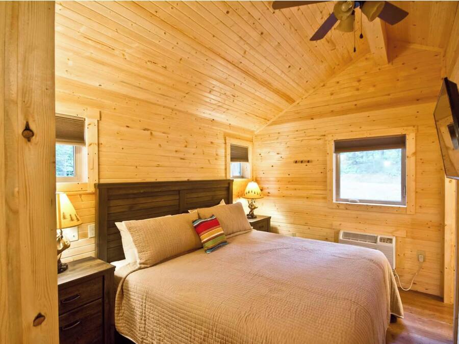 Un dormitorio acogedor de cabaña de madera con una cama, mesitas de noche, una ventana y un ventilador de techo.