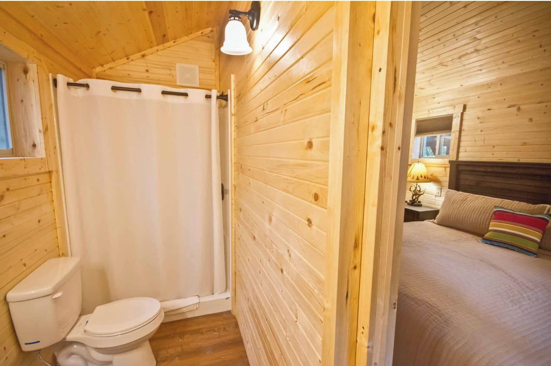 A wooden cabin with a bathroom featuring a toilet and a shower curtain, and a bedroom with a bed and a nightstand.