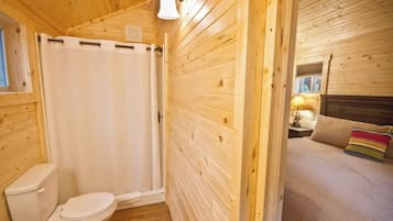 A wooden cabin with a bathroom featuring a toilet and a shower curtain, and a bedroom with a bed and a nightstand.