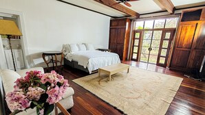 Un dormitorio con cama, piso de madera, ventilador de techo, ventana con vistas y mesita de noche con flores.