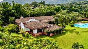 Una casa grande con piscina rodeada de exuberante vegetación y colinas al fondo.
