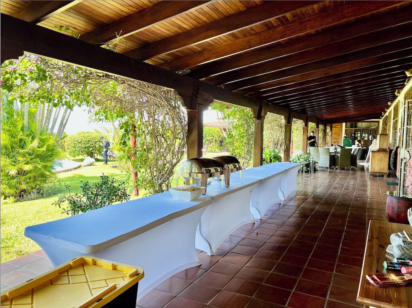 Un espace extérieur abrité comprenant une longue table blanche, un barbecue et une poubelle jaune.