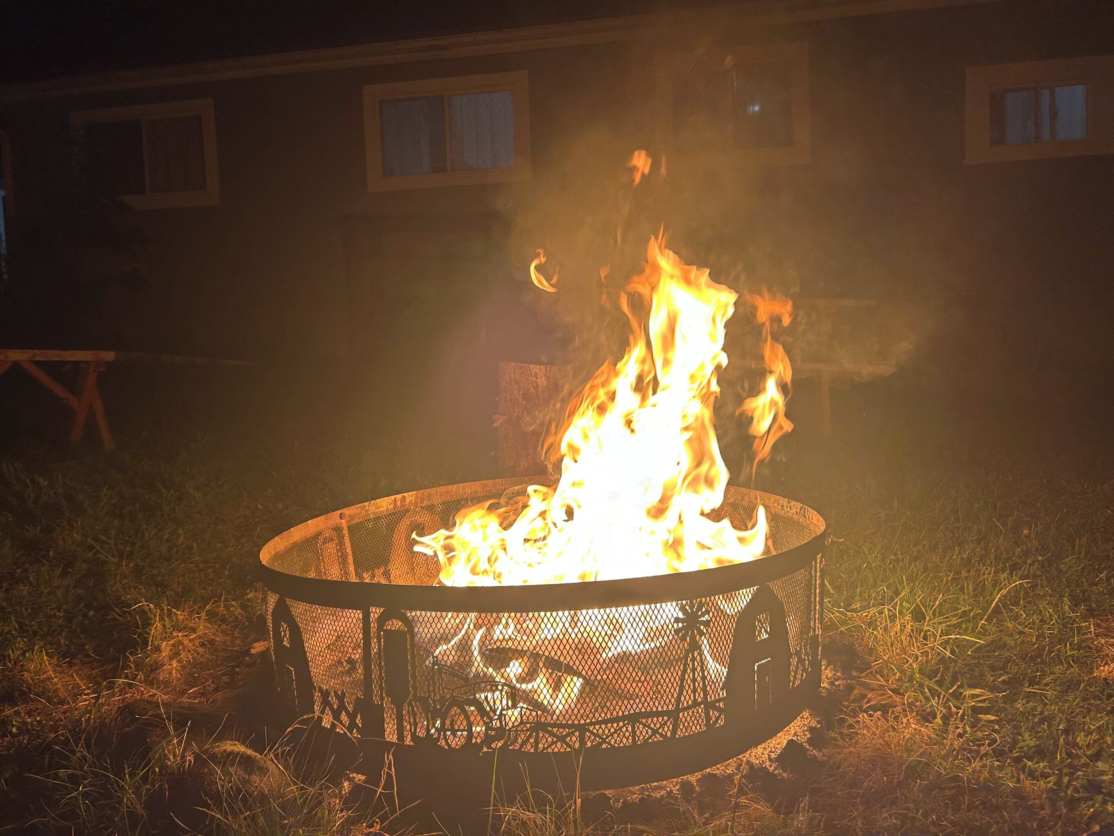 A metal fire pit with flames, surrounded by grass, with a building in the background.