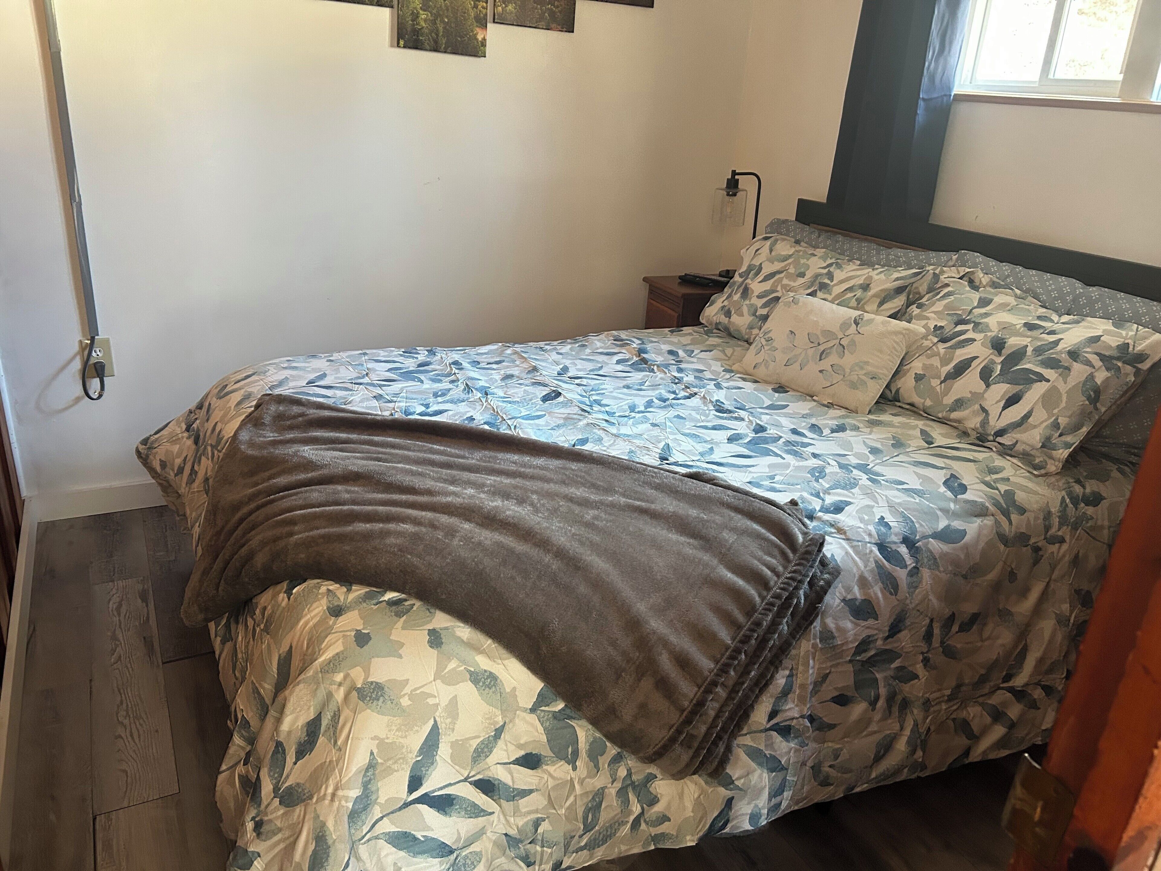A neatly made bed with floral bedding, a bedside table with a lamp, and a window with curtains.