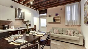 Una cocina moderna con techo de madera, un comedor con mesa dispuesta para cuatro personas, y una sala de estar con sofá y ventana con cortinas.
