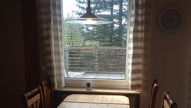 A dining area with a wooden table and chairs, a window with a view of a balcony and trees, and a clock on the wall. - Hexenstube Morgana Romantic apartment by the national park WLAN Pet-friendly, (Braunlage)