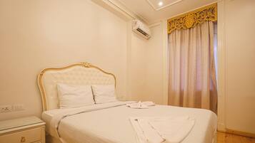A neatly made bed with white linens and a gold-trimmed headboard, a bedside table with a drawer, and a window with a decorative curtain.
