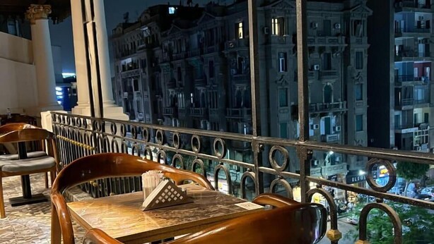 Een balkon met een houten stoel en tafel, met uitzicht op een stadsbeeld met historische gebouwen.