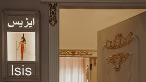 Een gang met een lijst waarop een afbeelding staat, het woord 'Isis' op de muur, een deur met decoratieve details en een stoel op de achtergrond.