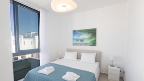 Habitación de hotel con cama, mesita de noche, un cuadro de una playa y vistas a edificios a través de un ventanal grande.
