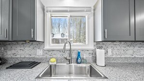 Cozinha com armários cinza, pia de aço inoxidável, janela com vista para casas e painel de fundo de mármore.